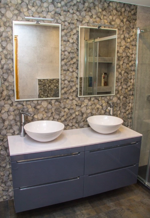 Reforma de baño con mueble suspendido gris, lavabos de bol y pared de piedra en Madrid